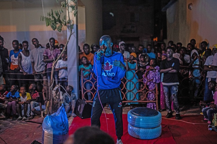 A man wearing a blue sweatshirt speaks into a microphone with a crowd surrounding him.