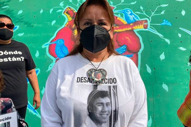 Letty, a woman with brown skin and brown hair, stares at the camera. She wears a black surgical mask and a t-shirt with her son Roy's image.