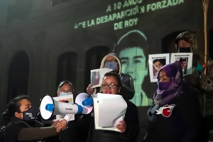 Letty uses a megaphone at a commemorative event. An image of her son Roy is displayed behind her. She is surrounded by four other people.