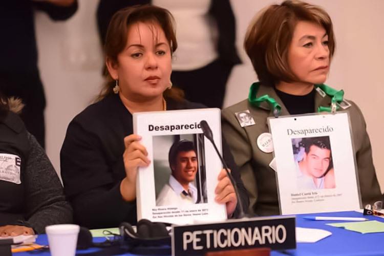 Letty and another woman, both with serious facial expressions, sit at a table holding photos of their missing sons.
