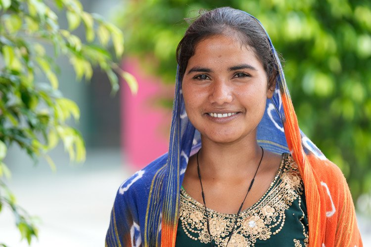 Meher, a young woman smiling and wearing a blue and orange scarf, was photographed at AMIED's workshop in Kishangarhbaas, India, August 2022. Photo by Deepak Sharma.