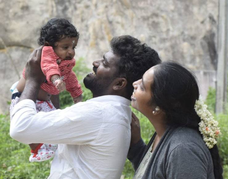 A young woman, Kavitha, wears flowers in her hair and stands with her husband Narendra, who holds up their baby boy. They are all smiling.