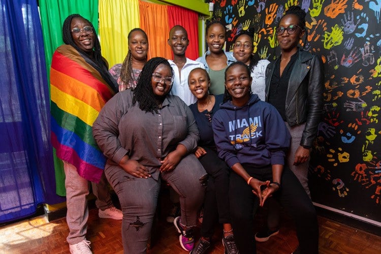 9 members of the NGLHRC team, all smiling and looking at the camera, gather in their Nairobi office — a bright, colorful space where they hold social events, workshops and offer legal guidance. There are colorful hand prints painted on a black wall and cutrains that form a rainbow flag behind the people in the photo. One person is wearing a rainbow flag as a cape.