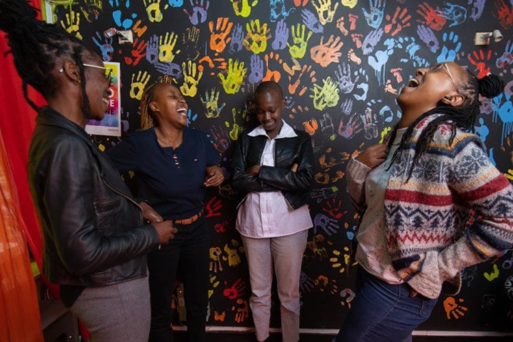 Four members of the NGLHRC team in front of a rainbow handprint wall, all laughing together. Photo by Lameck Ododo.