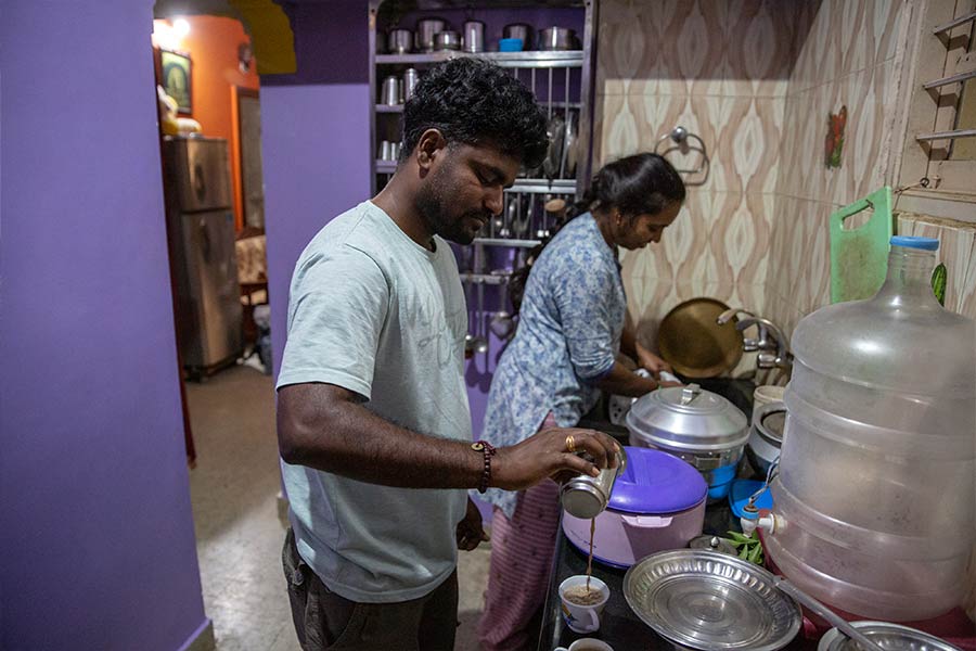 Narendra, a young man wearing a white t-shirt, pours tea into a cup in the kitchen with his wife Kavitha, who wears a floral top and is cooking.