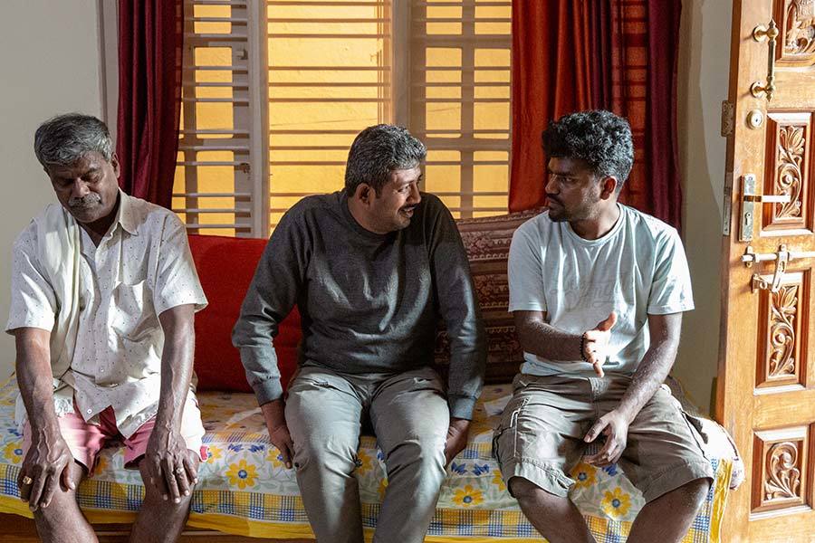 Three men sit on a sofa indoors speaking.