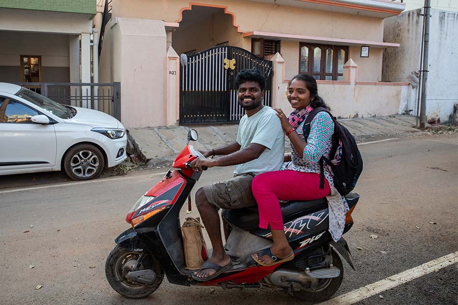 Narendra, a young man wearing a white t-shirt and brown shorts, sits on a motorbike with his wife Kavita, who wears a floral top and pink pants, giving her a ride to the train station for her daily commute.